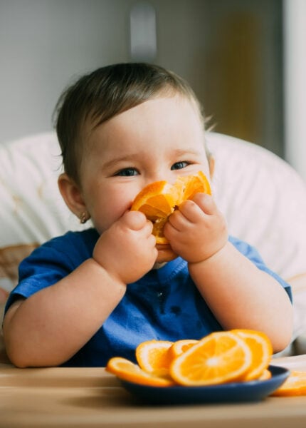 a little girl in a blue t-shirt and a blue plate sitting in a child's chair eating an orange