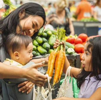 Holding her baby, a mother selects a bunch of carrots at the grocery store with her young daughter looking on